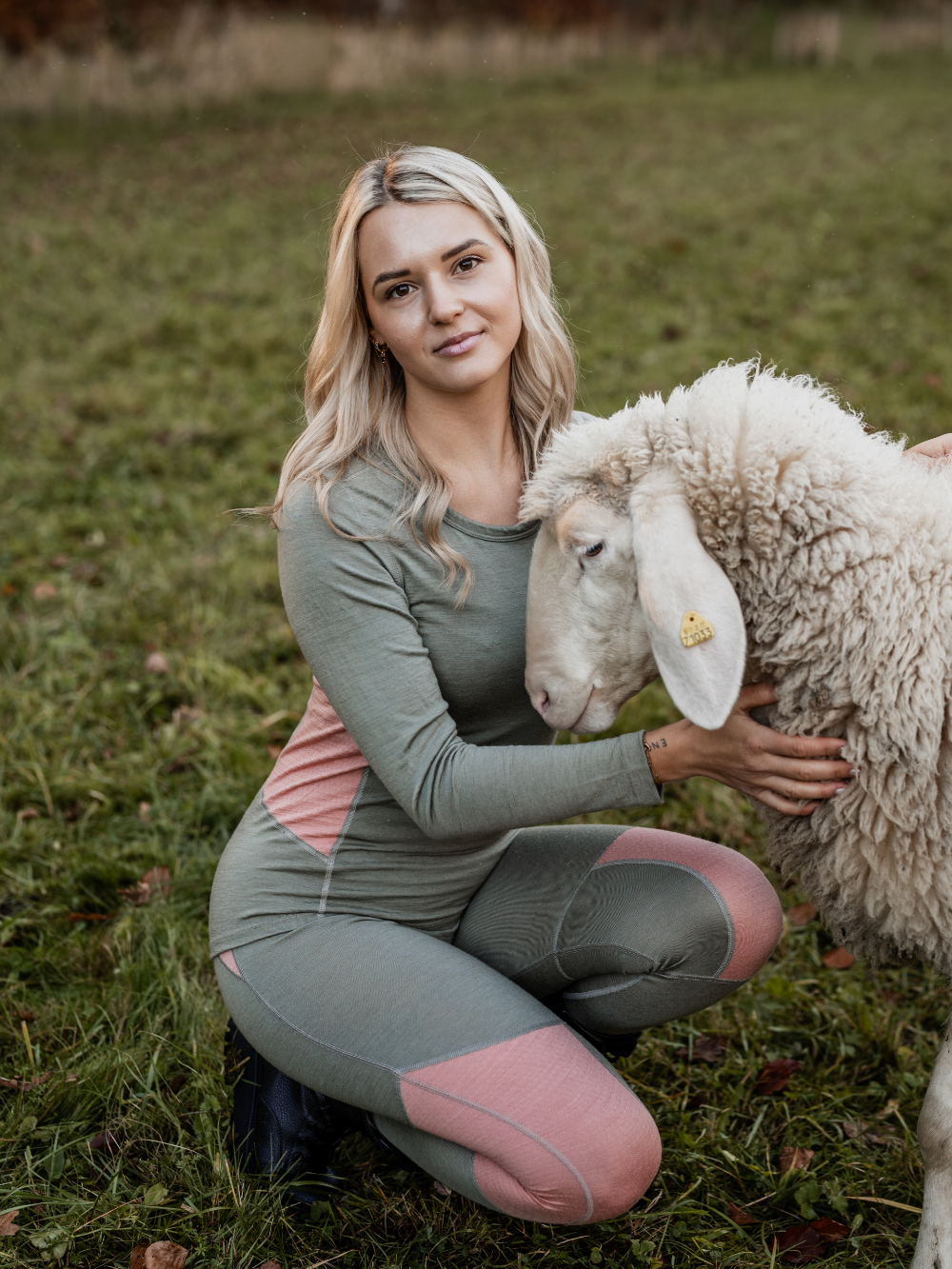 Frau in farbiger Merino-Funktionswäsche von Comazo kniet neben einem Schaf – natürliche Wärme und Komfort aus nachhaltiger Merinowolle.