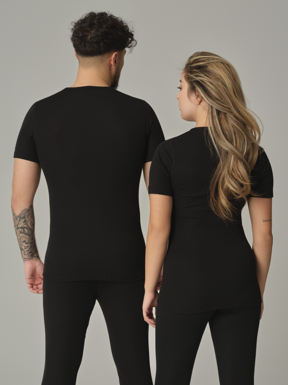 Rückansicht: Eine Frau und ein Mann tragen Unisex Funktionsshirt "warm extra" kurzarm von Comazo in der Farbe Schwarz mit angerauter Innenseite zur optimalen Wärmespeicherung, Feuchtigkeitsmanagement und verlängertem Rückenteil
