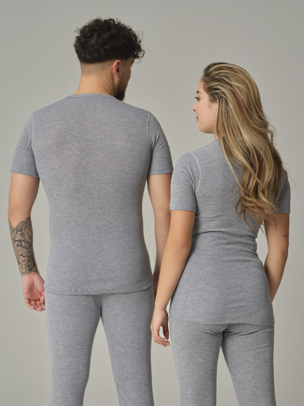 Rückansicht: Eine Frau und ein Mann tragen Unisex Funktionsshirt "warm extra" kurzarm von Comazo in der Farbe Grau-Melange mit angerauter Innenseite zur optimalen Wärmespeicherung, Feuchtigkeitsmanagement und verlängertem Rückenteil
