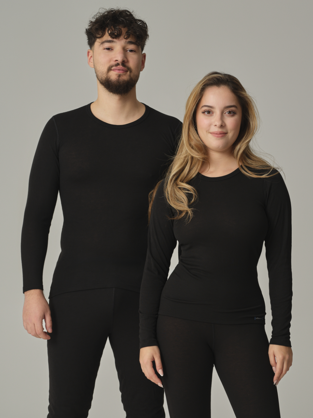 Vorderansicht: Eine Frau und ein Mann tragen Unisex Funktionsshirt "warm extra" langarm von Comazo in der Farbe Schwarz mit angerauter Innenseite zur optimalen Wärmespeicherung, Feuchtigkeitsmanagement und verlängertem Rückenteil