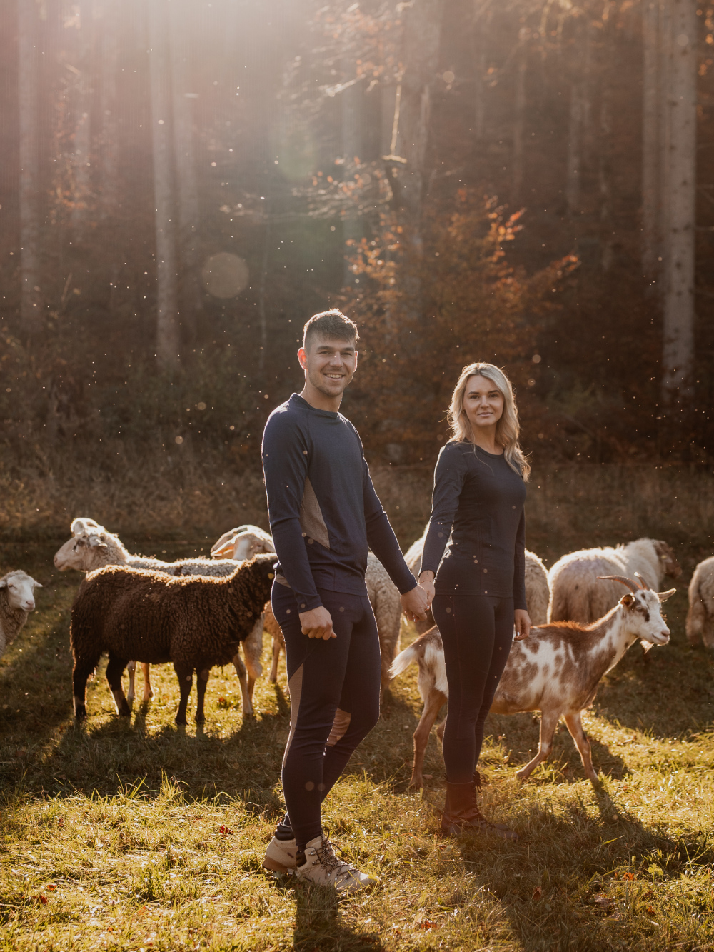 Paar trägt Merino-Funktionswäsche von Comazo und steht zwischen Schafen im warmen Abendlicht – natürliche Wärme und Atmungsaktivität aus Merinowolle